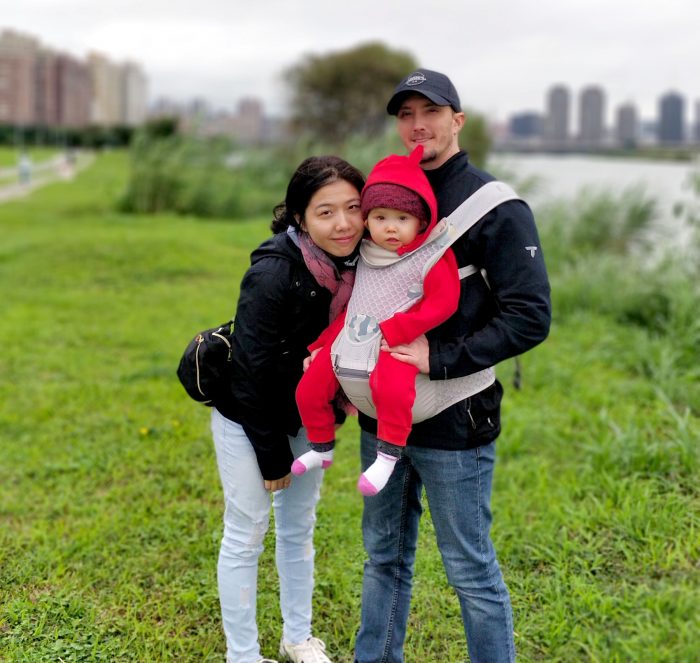Aspiration after feeding | AADC News | Richard holds Rylae-Ann in a hip seat baby carrier strapped around his body. Mom, Dad, and Rylae-Ann poses in an outdoor photo next to a river
