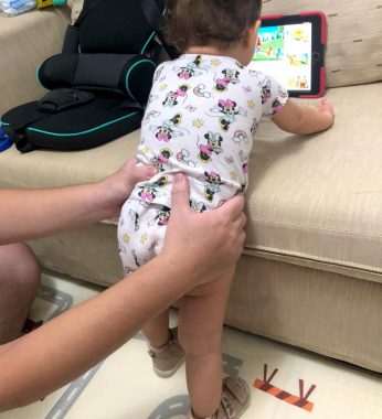 Standing with AADC | AADC News | Rylae-Ann stands and leans against the couch cushion to watch a show on an iPad. One of her parents holds her hips to support her.