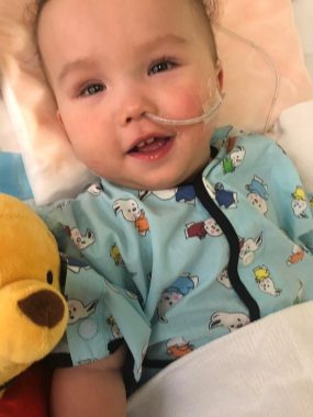 administering medication | AADC News | Rylae-Ann lies in a hospital bed with a feeding tube placed. She's wearing a hospital gown with bunnies and has a Winnie the Pooh stuffed toy next to her.