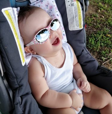 excessive sweating | AADC News | Rylae-Ann sits in her baby carrier in the shade under a tree at the park. She's wearing sunglasses and a white tank top.