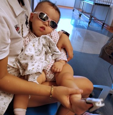 clinical trial | AADC News | Richard's wife sits in a hospital waiting room holding their daughter, Rylae-Ann, who is wearing sunglasses and has a pulse oximeter on her toe. 