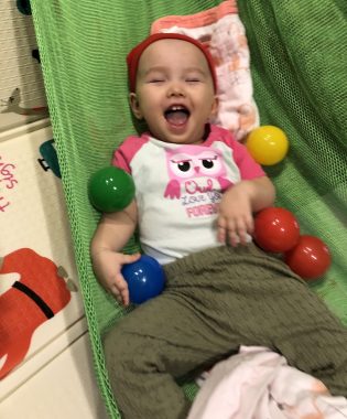 AADC symptoms | AADC News | Rylae-Ann, a member of the neurotransmitter disorder and rare disease community, laughs while lying on a green blanket and surrounded by colorful balls