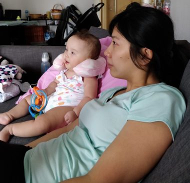 depression | AADC News | Rylae-Ann and her mom, Judy, sit on the sofa together. Rylae-Ann, pictured as a baby, is holding a colorful toy and has a supportive pillow behind her neck.