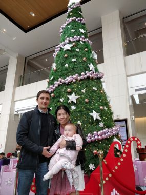 AADC deficiency diagnosis | AADC News | Richard, Judy, and Rylae-Ann pose in front of a large Christmas tree in what appears to be a mall in Taiwan in 2018.