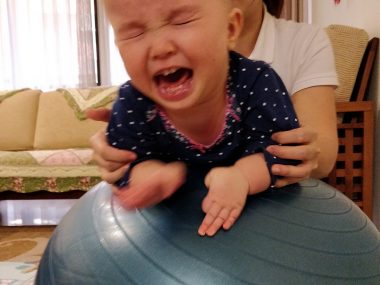 exercises | AADC News | Rylae-Ann facing the camera and crying while on a yoga ball, with adult hands holding her