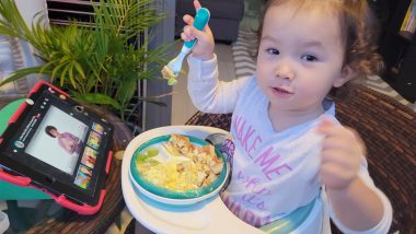 feeding issues | AADC News | Rylae-Ann uses a fork to eat independently. She is sitting in a highchair, with a plate of food and an iPad in front of her