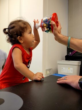 AADC deficiency | AADC News | During a physical therapy session, Richard holds a toy above Rylae-Ann's head, which captures her attention, while the physical therapist supports her during standing exercises.