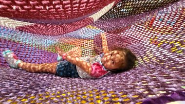 indoor playgrounds | AADC News | Rylae-Ann lies between large colorful nets at an indoor playground. the nets cast a patterned shadow across her.