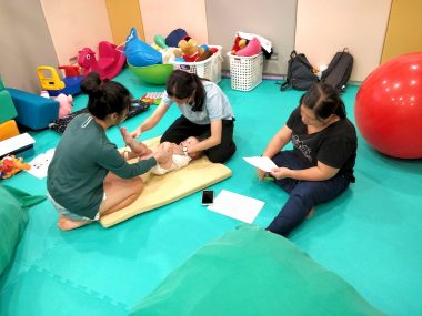 sensory integration therapy | AADC News | A trained therapist teaches Rylae-Ann's mom and nanny techniques they can use at home. The women are sitting on a turquoise mat surrounded by toys, and supporting Rylae-Ann as she lies on her back.