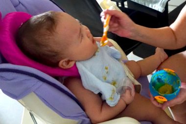 hypoglycemic crisis | AADC News | Rylae-Ann's parents feed her as she sits in a baby carrier. She wears a blue bib and is making an angry face