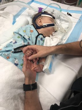 medical trauma | AADC News | Rylae-Ann, pictured as a baby, lies in a hospital bed as her father, Richard, holds her hands. She's hooked up to multiple tubes and machines.