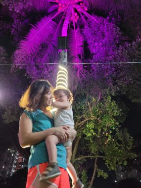 walking at night | AADC News | Richard's wife, Judy, holds their daughter, Rylae-Ann, during an evening walk. They are standing under a palm tree that's been lit up purple.