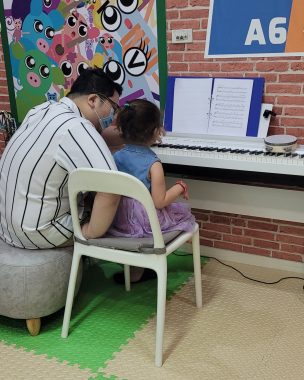 benefits of music therapy | AADC News | Rylae-Ann sits on a chair in front of a digital piano looking at sheet music. Her music therapist sits next to her. 