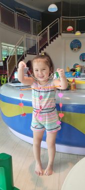 AADC deficiency | AADC News | Rylae-Ann smiles and holds up strings with pink hearts attached – an arts and crafts project she made in class.