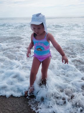 negative comments | AADC News | Rylae-Ann wades in the surf on the beach. She's wearing a pink and blue swimsuit and a hat.