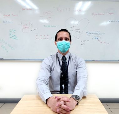 caregiver self-care | AADC News | Richard sits in a classroom in front of a white board filled with kind messages from his students. He is dressed in a shirt and tie, and wearing a mask, and has his hands casually clasped in front of him on a desktop