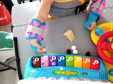 negative comments | AADC News | Rylae-Ann plays with several multicolored toys while being supported by her standing frame and wearing wrist braces.