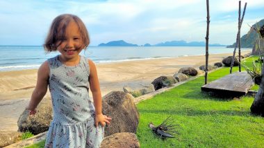 Rylae-Ann stands next to rocks near the beach, with the silhouettes of mountains looming in the background. She has short brown hair and is wearing a blue patterned dress and smiling.
