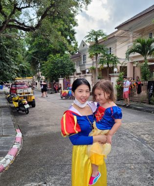A woman wearing a yellow skirt and a blue blouse with red and yellow sleeves stands in the middle of a residential street holding a toddler, who also wears a yellow skirt and a blue blouse with red in the center. There are what appear to be palm trees on one side near houses and greenery from another tree on the other side of the street.