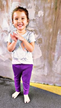 A standing Rylae-Ann smiles in front of a gray wall with some white and brown splotches. She wears purple pants and a very light blue/whitish T-shirt.