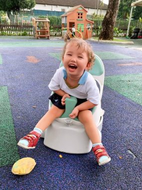 Rylae-Ann is seated on a small white chair while in an outdoor park of what appears to be artificial turf. She is smiling and wears a white shirt with a blue collar and red sandals. A yellow toy lies in front of her, with a playhouse in the distance behind her..