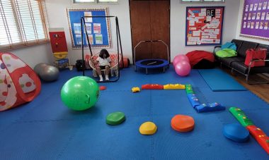 A photo of an inclusive classroom features blue mats on the floor, a swing, a child's trampoline, a small tent, and balls and toys in various sizes and colors.