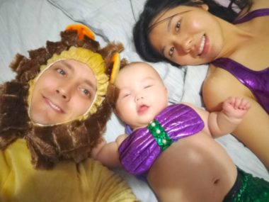 A man and woman lie on a bed with an infant between them. We see their faces. The man's is costumed with a yellow hood with what's designed to look like brown hair. The woman wears a purple tank top. The infant, whose torso we see, has her chest costumed in purple with green in the middle.