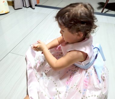 In a side view from above, a little girl sits on the floor in a pink and white dress belted with blue.