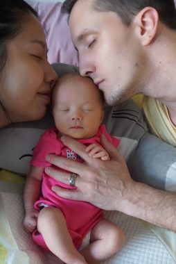 benefits of snuggling | AADC News | aadc deficiency | photo showing the infant Rylae-Ann, in a pink outfit, with the faces of Judy on one side and Richard on the other.