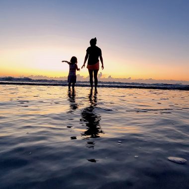 A photo depicts the silhouette of a woman and her young daughter walking on the beach at sunset. They are standing in shallow water, and the photo is taken at a low angle, making it appear as though they're walking on water. The woman is wearing a T-shirt and pink shorts, and the young girl is pointing toward the horizon.
