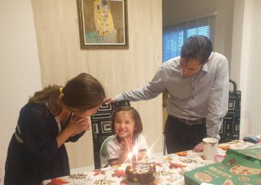 Two parents - a man and a woman - dote on a young child who sits at a table in front of a birthday cake with candles and smiles while clapping her hands