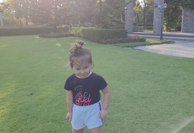 This photo shows a little girl standing in a broad, green lawn with well-manicured shrubs in the background. She has light brown hair tied up in a bun and wears a dark blue shirt with light blue shorts.