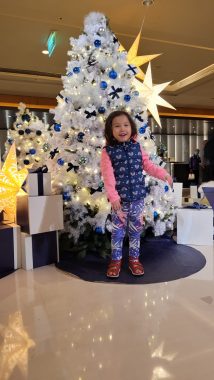 This photo shows a young girl standing in front of a small white Christmas tree decorated with blue ornaments. The tree is surrounded by white boxes, meant to look like presents, and large, lit-up stars. The girl is smiling and wearing a long-sleeve pink shirt, blue vest, and purple patterned leggings.