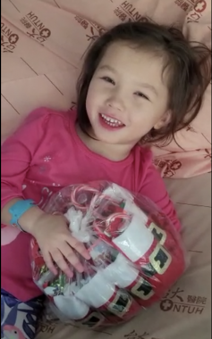 This photo shows a young girl lying down and holding a bag full of small stockings containing goodies for other pediatric patients in the hospital. She is smiling, wearing a pink shirt, and has a blue hospital bracelet on her wrist.