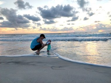 A young father crouches down in the surf at a beach in Florida. He's holding his 3-month-old daughter so that her feet touch the water. The ocean looks calm, and the sun is setting in the background.