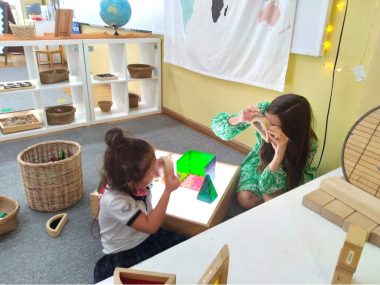 A young girl sits on the floor in her classroom next to her teacher. In between them is an illuminated box with toys of various shapes and colors on top of it. The girl and her teacher are both holding up wooden half circles and looking at each other through the whole in the center. In the background are shelves with other activities, a globe, a whiteboard, and a map of the world.