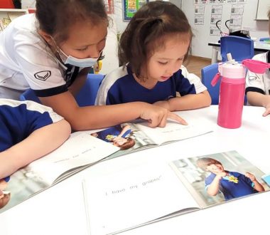 A young girl sits at a round table with a book open in front of her. Other students sit on either side of her, with their books open to the same page. A person wearing a face mask stands behind the girl, pointing to the words on the page.