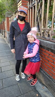 This photo shows a woman and a young girl standing on a sidewalk next to a brick wall. They are both dressed in warm clothes, with the woman wearing a long gray coat and colorful hat and the girl wearing a puffy pink and purple jacket, red skirt, black leggings, and a pink pom-pom hat.
