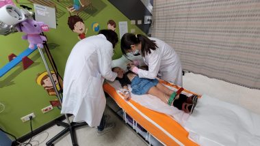This photo shows a young girl lying on her back in a pediatric hospital bed as two nurses in white coats attend to her and conduct various tests. The wall behind them is green and depicts illustrations of children.