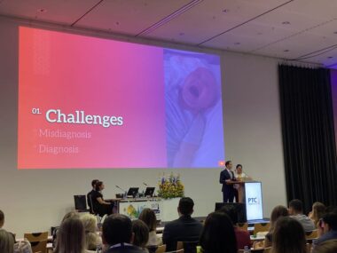 A mid-distance photograph shows two speakers behind a podium in a conference hall full of people. A projection on a screen behind the speakers shows the word "challenges." 