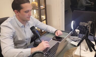 A young man in a formal dress shirt sits behind a laptop computer and a microphone, with a light projected at him - all indications that he is recording a podcast.