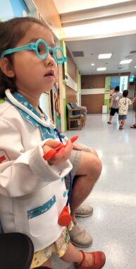 A young girl stands in a hallway in the children's ward at the hospital. She's wearing a doctor costume, including green plastic glasses, a white coat, and a stethoscope around her neck. Other children are visible in the background walking down the hall.