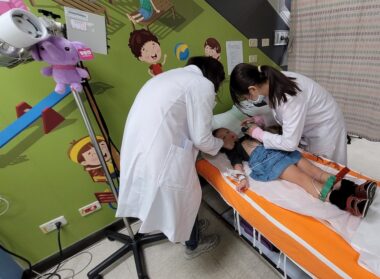 A young girl lies on a hospital bed as two medical providers in white coats and face masks attend to her. They appear to be in the pediatric unit of the hospital, as the bed is bright orange, the wall is green and features a painting of children, and there's a purple stuffed animal attached to a piece of medical equipment.