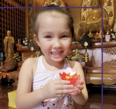 A young girl smiles while holding a cup filled with something red. Her hair is tied back with a gold bow and she's wearing a pink tank top. She appears to be inside a religious temple, as there are various statues of monks behind her.