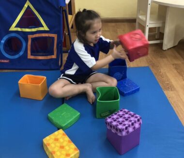 A young girl sits on a blue mat with large blocks of various colors and shapes.