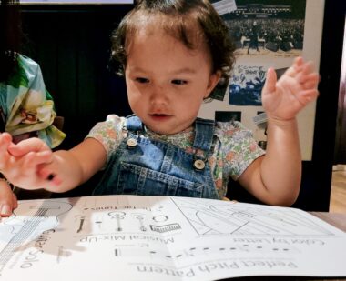 A young girl, perhaps a toddler still, sits at a table with a large educational book of drawings before her. She has her arms raised and looks down at the book. 