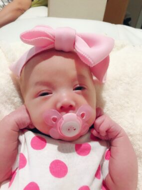 A close-up photo of a newborn baby girl with a pink bow on her head, a pink pacifier, and a white and pink polka dot onesie. 