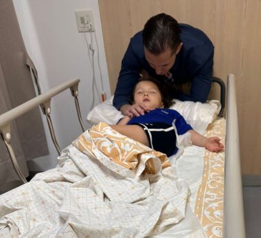 A 5-year-old girl lies on a hospital bed after breaking her arm on the playground. Her father stands behind her with his hand on her shoulder.