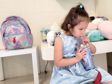 A young girl, in a light blue dress with small, largely white flowers, sips from a straw in a large, cupped drink. Behind her at left is a multicolored backpack on a white table. Behind her at right is another white table with stuffed animals on top.