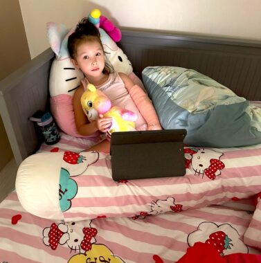 A 5-year-old girl sits propped up in bed with her left arm in a pink cast. She's surrounded by stuffed animals and is watching videos on a tablet. She looks directly at the camera. 